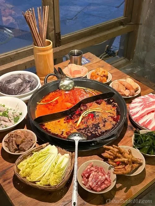 A bubbling dual-flavor spicy Sichuan hotpot surrounded by various side dishes including sliced beef, tripe, goose intestines, and fresh vegetables on a wooden table.