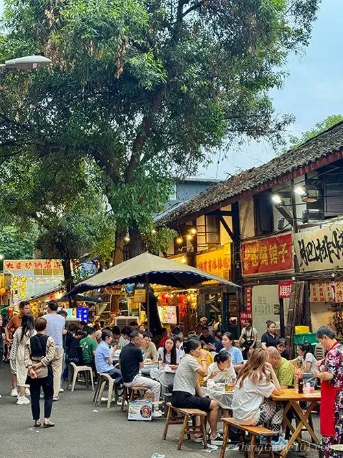 A bustling outdoor 'Fly Restaurant' scene in a Chengdu alley, with many local people sitting on small stools at simple tables under trees and umbrellas, surrounded by traditional buildings and food stalls.
