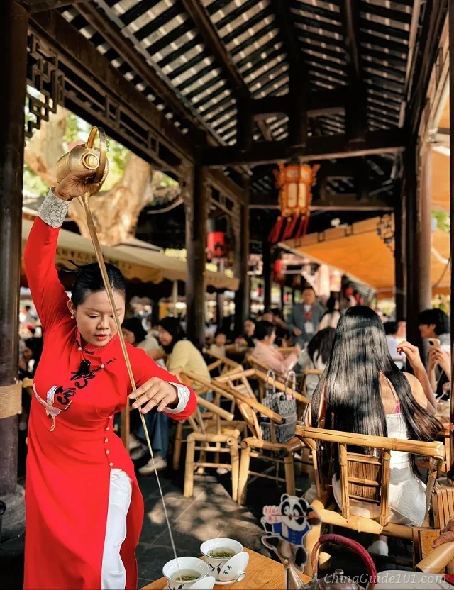 A tea master in a traditional red dress performing the long-spout teapot ceremony in a crowded open-air Chengdu teahouse.