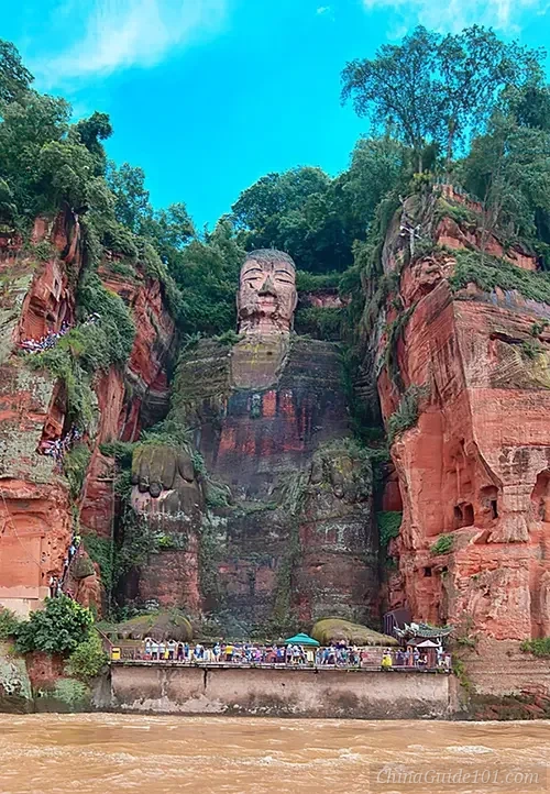 The Leshan Giant Buddha, a massive 71-meter tall stone statue carved into a red sandstone cliff overlooking the river, with tourists visible at its base for scale.