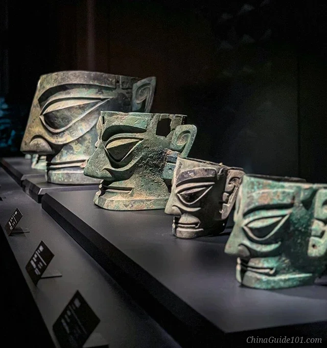 A row of ancient bronze masks with distinctive almond-shaped eyes and large ears displayed on black pedestals inside the Sanxingdui Museum.
