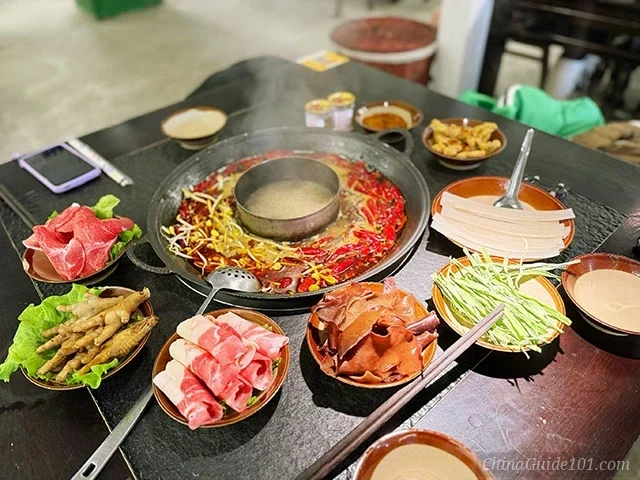 A traditional Chongqing hotpot set on a black table, featuring a split pot with spicy red broth full of dried chilies and a clear center broth, surrounded by fresh ingredients like sliced beef, chicken feet, wood ear mushrooms, and greens.