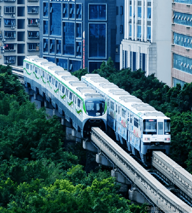 Two Chongqing Rail Transit Line 2 monorail trains passing each other on elevated tracks, surrounded by lush green trees with modern blue-glass residential buildings in the background.