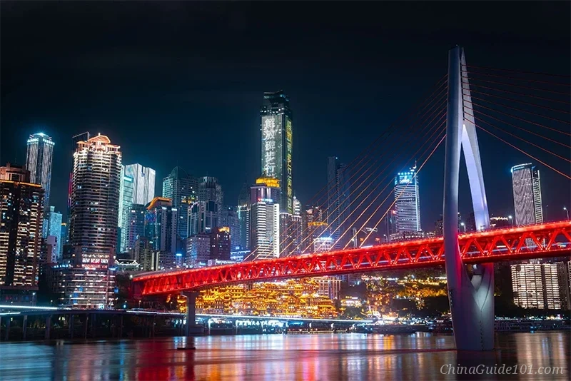 A stunning night view of Chongqing's cyberpunk skyline featuring the illuminated Hongyadong stilt houses, modern skyscrapers, and the red-lit Qiansimen Bridge spanning the Jialing River.