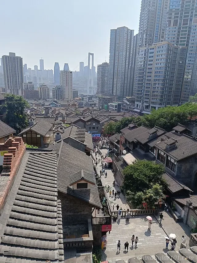 A high-angle view of Shibati Traditional Old Street in Chongqing, showing grey-tiled rooftops of traditional buildings contrasted against a backdrop of modern skyscrapers and the Caiyuanba Bridge.
