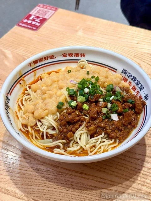 A bowl of authentic Chongqing Wanza Mian, featuring alkaline wheat noodles topped with creamy mashed yellow peas, savory minced pork sauce (Zajiang), and chopped green onions in a spicy chili oil base.