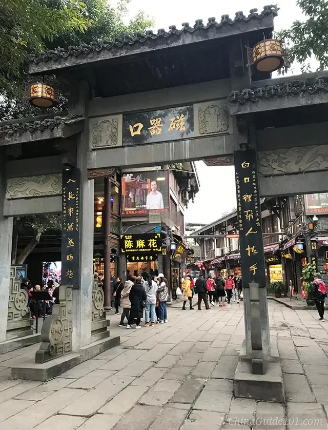 The grand stone entrance gate of Ciqikou Ancient Town in Chongqing, featuring traditional calligraphy and lanterns, with a bustling street of shops and visitors in the background.