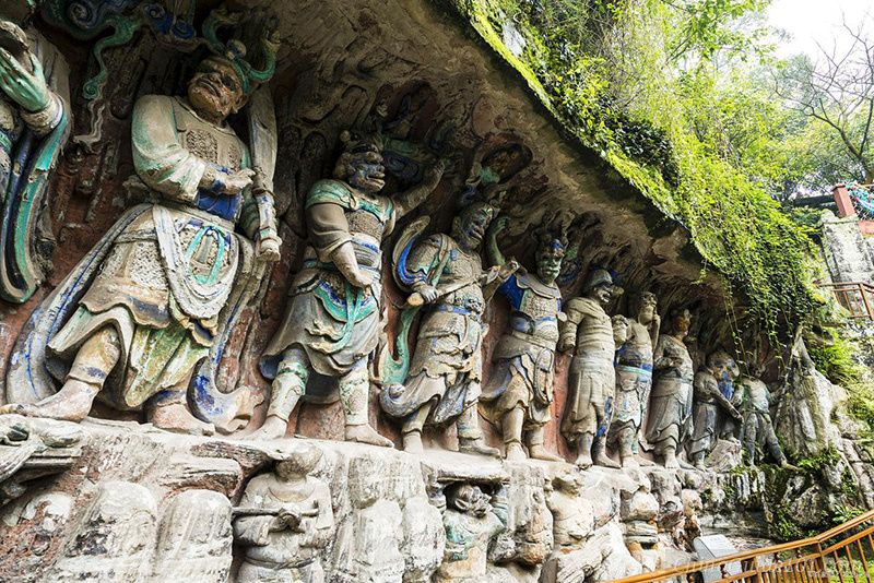 Intricately carved and colorful Buddhist stone statues of Guardian Deities at Baoding Mountain, Dazu Rock Carvings. The relief features multiple towering figures in traditional armor with expressive faces, carved directly into the limestone cliffside under a mossy overhang.