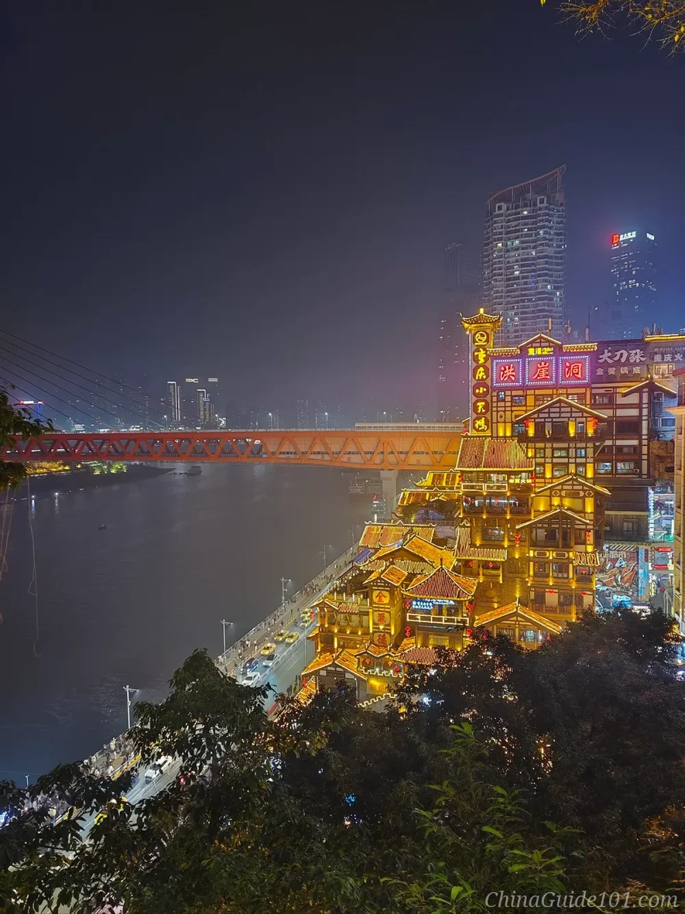 Night view of Hongyadong in Chongqing, featuring a multi-story traditional stilt house complex glowing with golden lights next to the Jialing River and the red Qiansimen Bridge.