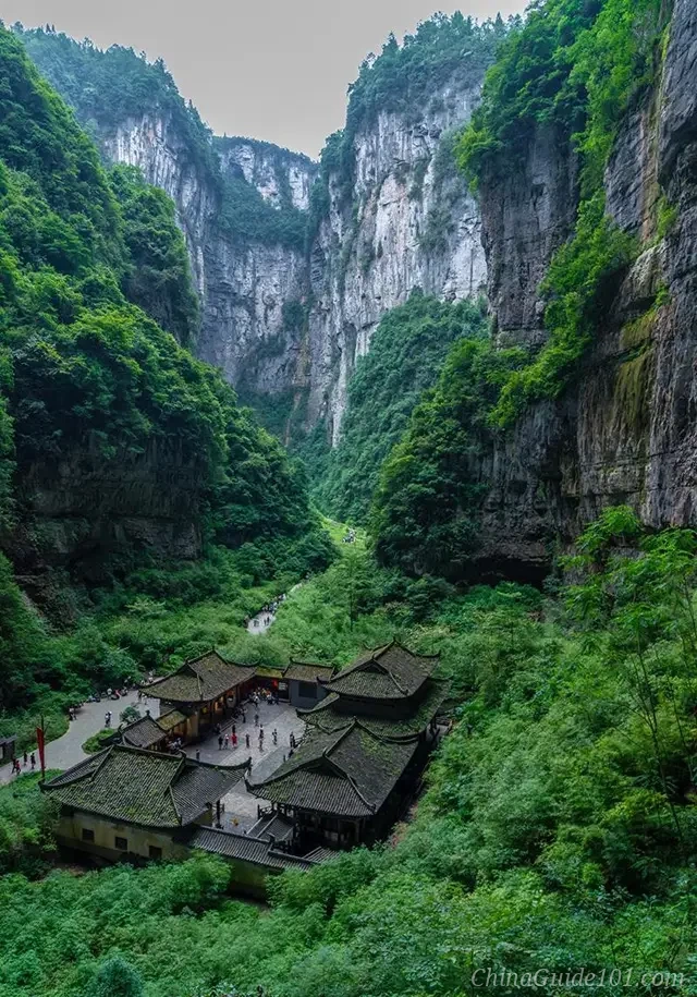 A wide-angle view of the Tianlong Post House, an ancient-style Chinese courtyard building with dark tiled roofs, nestled at the bottom of a deep limestone gorge in Wulong Karst National Park, surrounded by towering vertical cliffs and lush greenery.