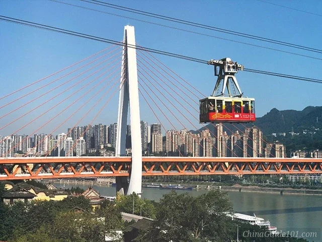 A red and beige Yangtze River Cableway car gliding over the river, with the white pylons and orange trusses of the Dongshuimen Bridge and the city skyline in the background.