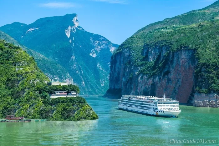 A large multi-deck white luxury cruise ship sailing through the turquoise waters of the Qutang Gorge on the Yangtze River, flanked by towering vertical limestone cliffs and lush green mountains under a clear blue sky.