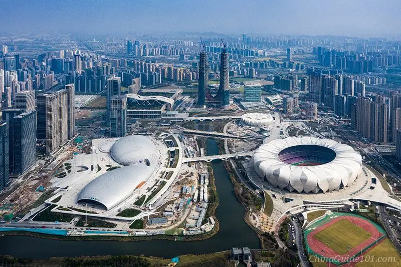 An aerial wide-angle view of the Hangzhou Olympic Sports Center, featuring the iconic white lotus-shaped main stadium and the butterfly-shaped sports and swimming pool complex. The futuristic structures are surrounded by a canal and modern skyscrapers under a clear blue sky.