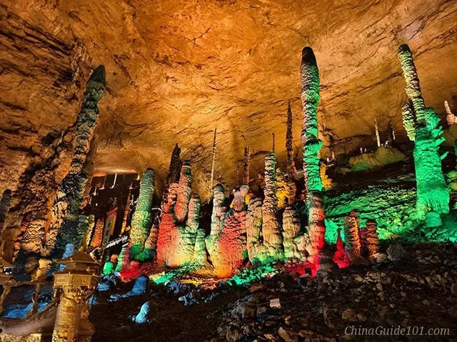 The interior of Huanglong Cave in Zhangjiajie featuring numerous tall, slender stalagmites rising from the cavern floor. The rock formations are dramatically illuminated with vibrant green, red, and yellow artificial lights against the natural earthy tones of the cave ceiling