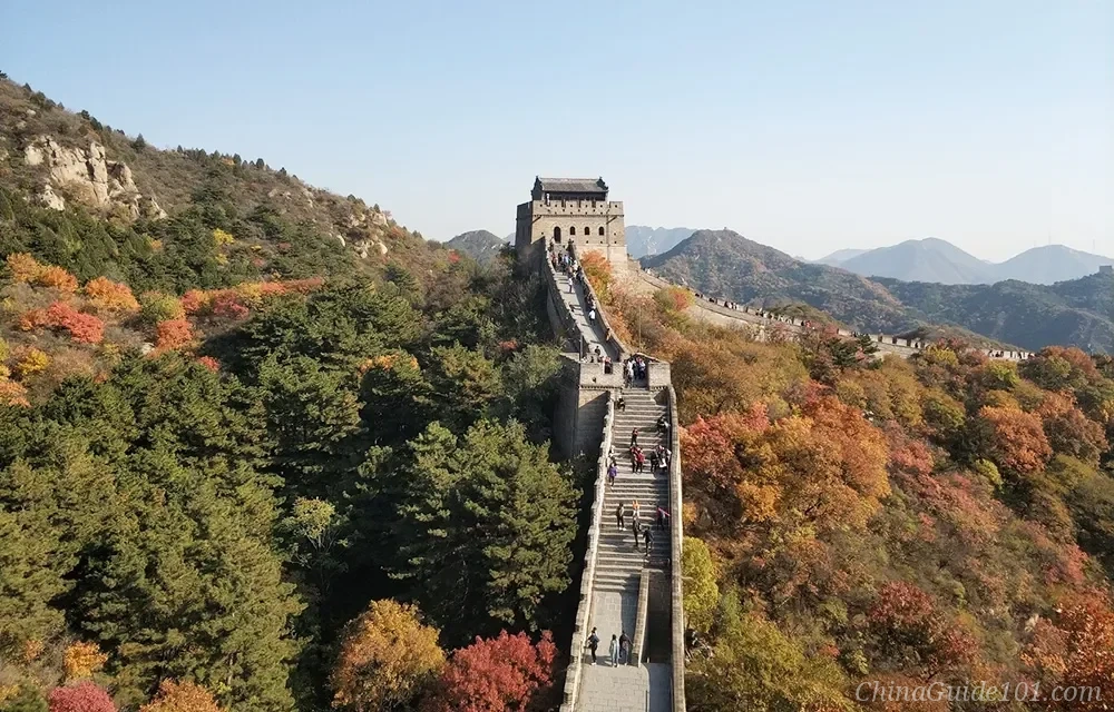 Tourists climbing the steep stone steps toward a prominent two-story watchtower at Badaling Great Wall in Beijing, surrounded by vibrant autumn foliage with red and yellow leaves under a clear sky.