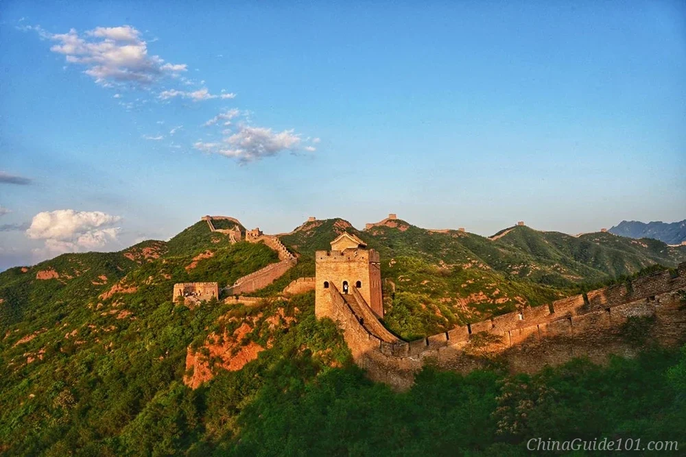 The Jinshanling Great Wall snaking across vibrant green mountain ridges during the golden hour sunset, featuring a prominent ancient watchtower bathed in warm orange light under a blue sky.
