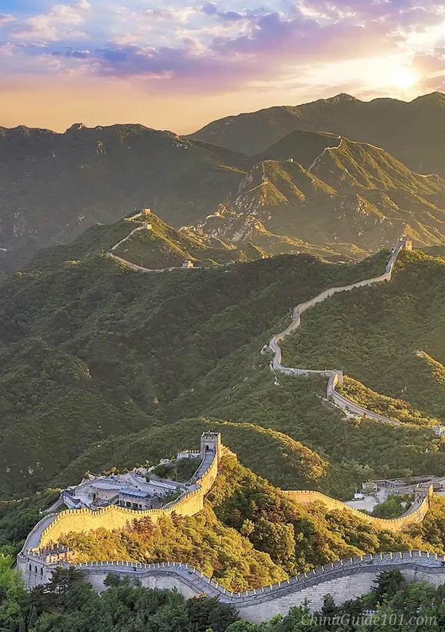 An aerial panoramic view of the Mutianyu Great Wall at sunset, showing the famous Zhengguan Terrace in the foreground and the stone wall snaking in an S-shape across lush green mountain ridges under a dramatic golden and purple sky