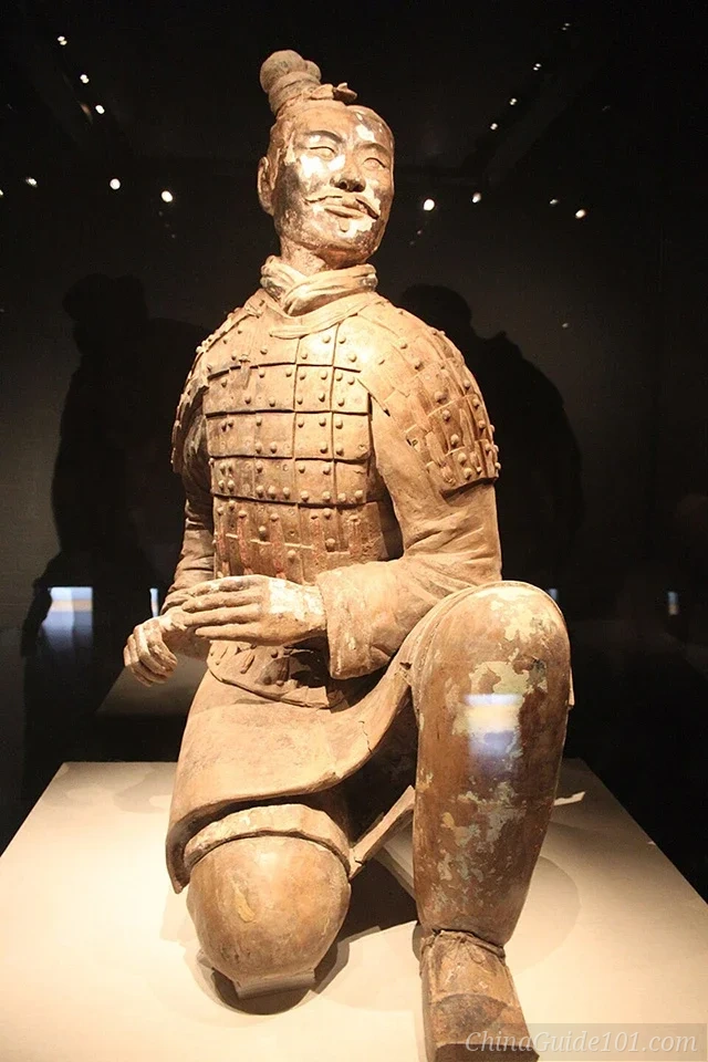 A close-up shot of an ancient Chinese terracotta kneeling archer statue on a museum display stand, wearing riveted armor with a topknot hairstyle, kneeling on his right knee while readying his hands to hold a crossbow.
