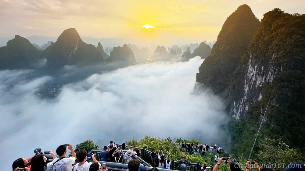 Panoramic sunrise view from a high vantage point overlooking the sea of clouds and karst mountain peaks in Yangshuo. The bright golden sun breaks through the sky, casting warm light on a thick blanket of low-lying fog and mist filling the valley below. The tops of tourists and a viewing platform are slightly visible at the bottom edge.