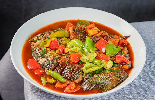 A close-up shot of Yangshuo beer fish served in a large white ceramic bowl. The dish features a pan-fried whole fish with scored skin, simmered in a rich, reddish-brown savory sauce, and generously topped with chunks of red and green bell peppers, chopped tomatoes, and sprinkled with fresh green scallions.