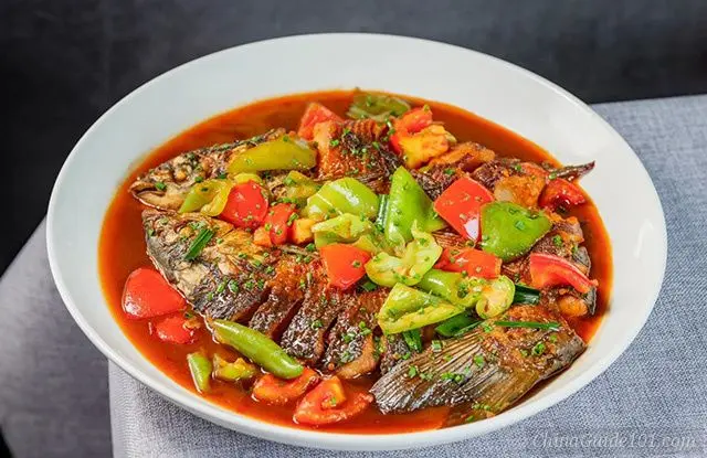 A close-up shot of Yangshuo beer fish served in a large white ceramic bowl. The dish features a pan-fried whole fish with scored skin, simmered in a rich, reddish-brown savory sauce, and generously topped with chunks of red and green bell peppers, chopped tomatoes, and sprinkled with fresh green scallions.