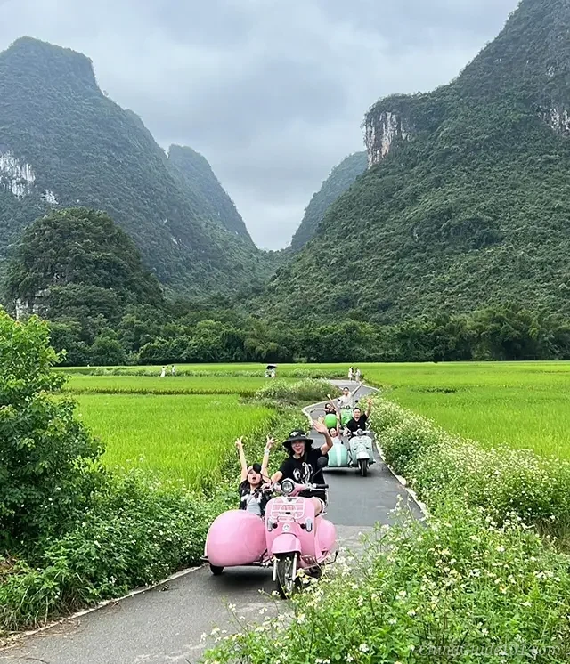 A group of cheerful tourists riding colorful vintage-style electric scooters with sidecars down a narrow paved path. The path cuts through lush vibrant green rice fields, with spectacular karst mountain peaks rising under an overcast sky in Yangshuo.