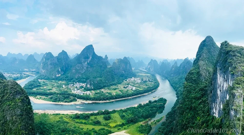Panoramic high-angle view of the Li River curving through lush green karst mountain peaks in Yangshuo, Guilin, under a cloudy blue sky.