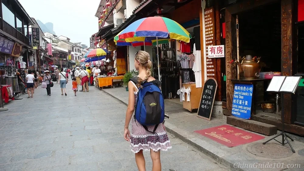 A blonde female tourist wearing a backpack, walking through the West Street pedestrian area in Yangshuo. The street is lined with traditional Chinese buildings housing souvenir shops, cafes, and bars with signs in both English and Chinese