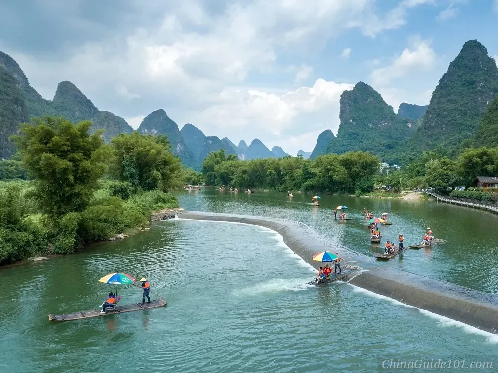 Traditional bamboo rafts with colorful umbrellas cruising down the Yulong River in Yangshuo, Guilin. Rafts are passing over a low concrete weir or dam, surrounded by lush green bamboo trees and dramatic karst mountain peaks under a cloudy sky.