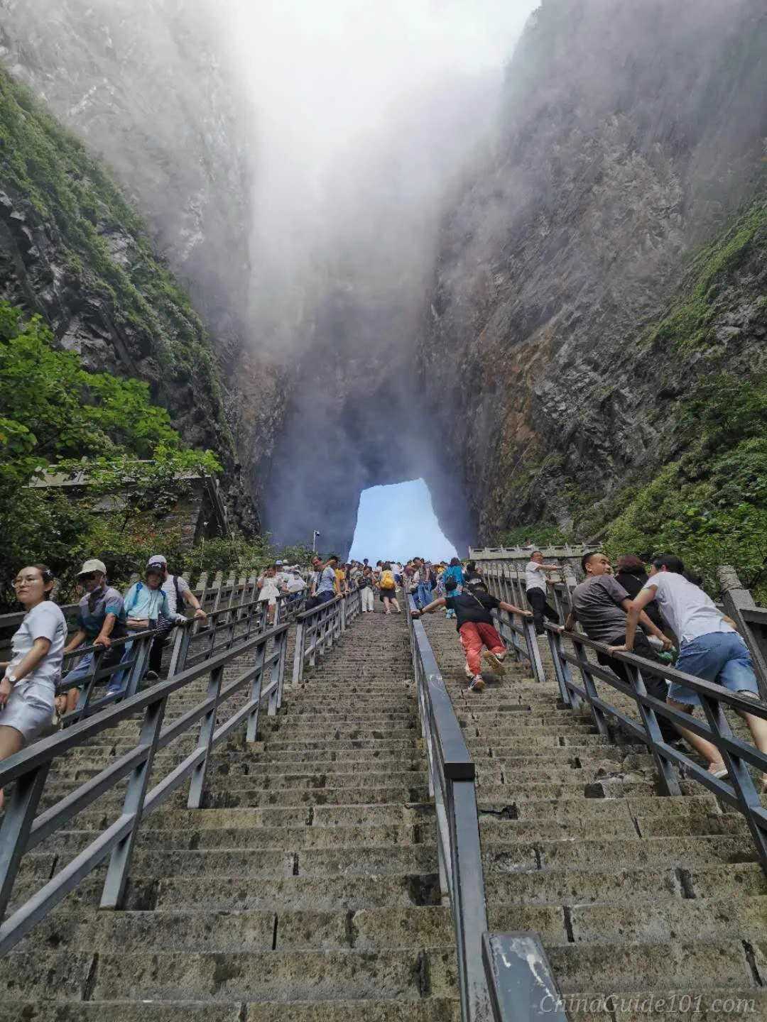 A low-angle, first-person perspective looking up a very steep stone staircase with handrails, where numerous tourists are climbing toward a massive natural rock archway. Heavy white mist and clouds swirl between the towering vertical cliffs, partially obscuring the top of the mountain.