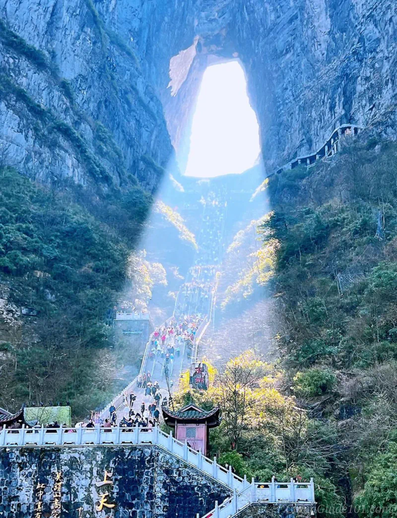 A distant, wide landscape view of a massive mountain range with a prominent, natural circular hole piercing through the peak, known as Heaven's Gate. A winding mountain road with sharp switchbacks curves dramatically down the lush green valley in the foreground under a bright sky.