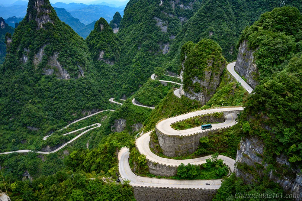 A high-angle aerial view of a winding mountain highway with multiple sharp, sweeping hairpin turns carved into steep, lush green mountain peaks. A small blue shuttle bus is driving along one of the elevated concrete curves built against the stone cliffs.