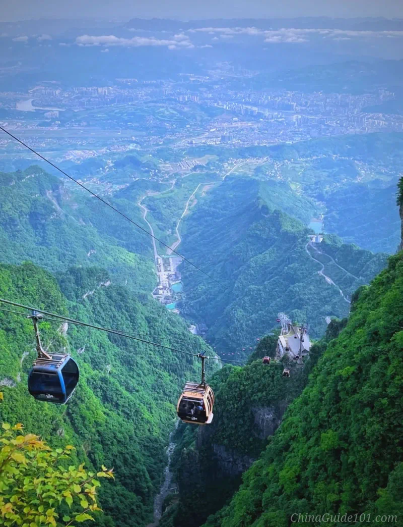 A red cable car cabin moving along a steel cable line, suspended high above steep mountain slopes and deep green valleys covered in thick trees and misty forests under a slightly cloudy sky.