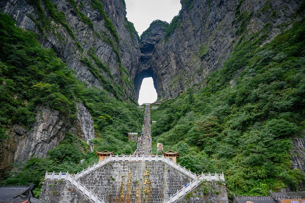 A massive, natural limestone archway known as Heaven's Gate on Tianmen Mountain, with a steep stone staircase featuring 999 steps leading up to the opening. Green vegetation covers the surrounding sheer vertical cliffs under a soft, slightly overcast sky