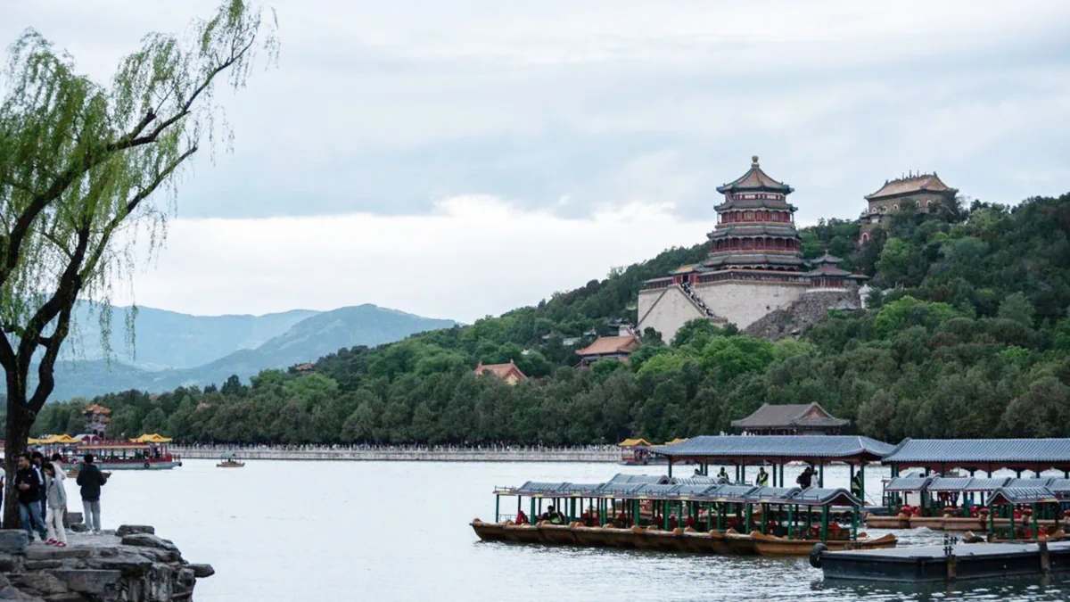 Scenic panoramic view of Kunming Lake with traditional dragon boats, framed by green trees, with Longevity Hill and the Tower of Buddhist Incense rising in the background at the Summer Palace in Beijing.