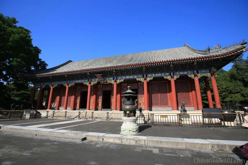 Wide shot of the Hall of Benevolence and Longevity at the Summer Palace, featuring traditional red pillars, grey tiled roofs, and a bronze incense burner in the courtyard under a clear blue sky.