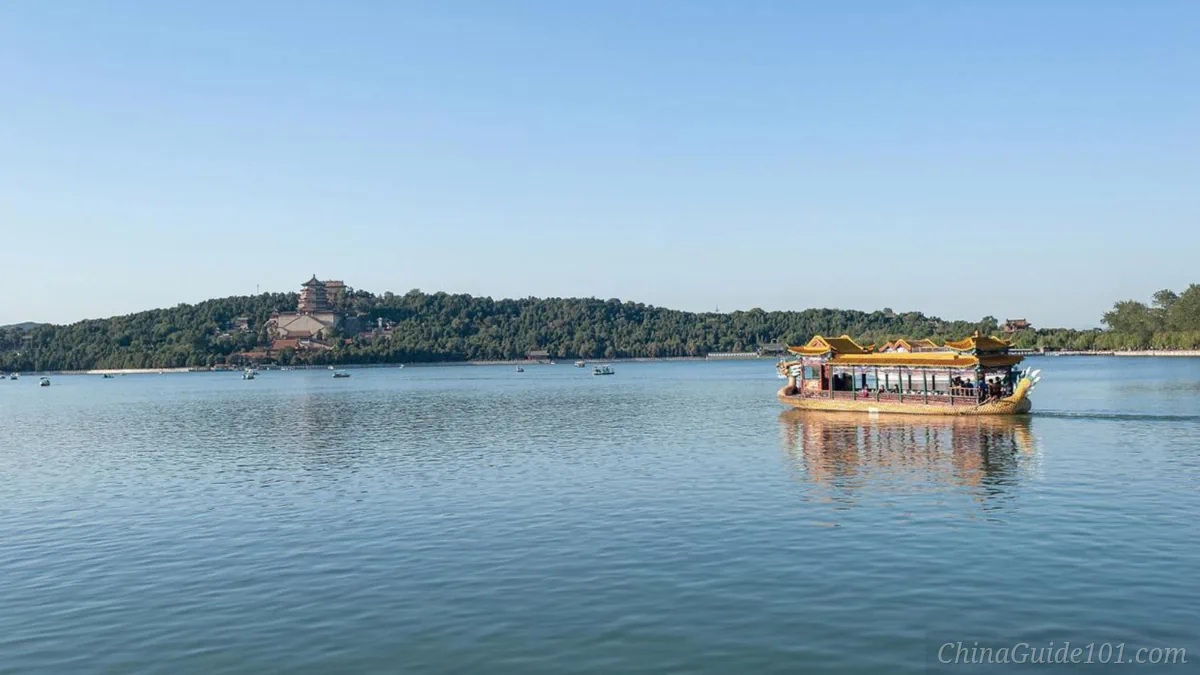 A traditional Chinese dragon boat cruising on the calm blue waters of Kunming Lake, with Longevity Hill and the Tower of Buddhist Incense visible across the water under a clear blue sky.