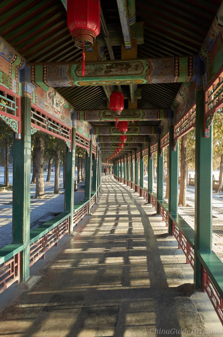 Interior perspective view of the covered wooden Long Corridor at the Summer Palace, decorated with traditional Chinese paintings, red lanterns, and sunlight casting long shadows on the walkway.