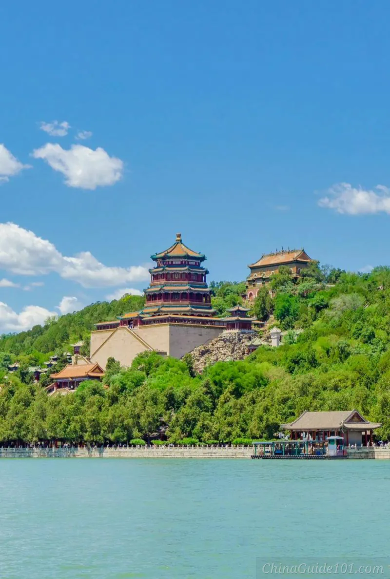 Vertical view of the Tower of Buddhist Incense standing tall on lush green Longevity Hill, overlooking the waters of Kunming Lake under a bright blue sky with white clouds.