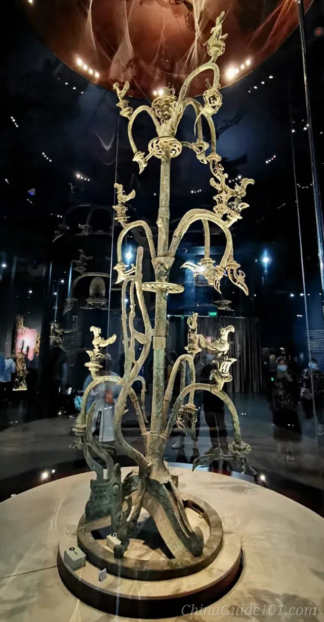 Full view of the towering ancient Sanxingdui Bronze Sacred Tree in a circular glass showcase, featuring intricate branches adorned with birds and flowers under museum spotlights.