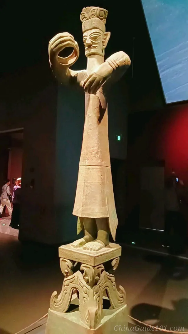 Full-length view of the towering ancient Sanxingdui Bronze Standing Figure on its elaborate pedestal, displayed under spotlights inside the museum hall, showing its oversized hands and intricate robes.