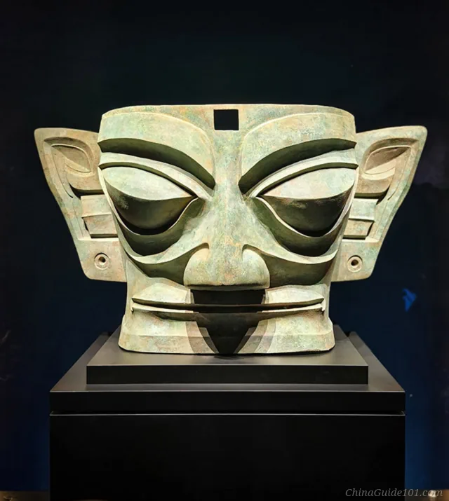Front view of a large ancient Sanxingdui bronze mask with exaggerated almond eyes, wide ears, and a prominent nose on display against a dark background.