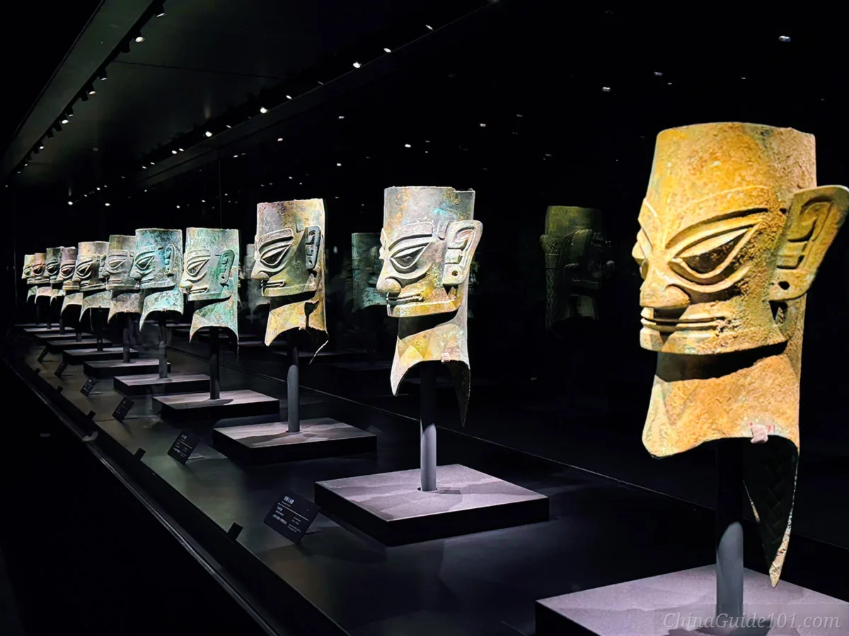 A long row of ancient ancient bronze heads and gold-masked bronze heads displayed on pedestals inside a dark exhibition hall at Sanxingdui Museum.