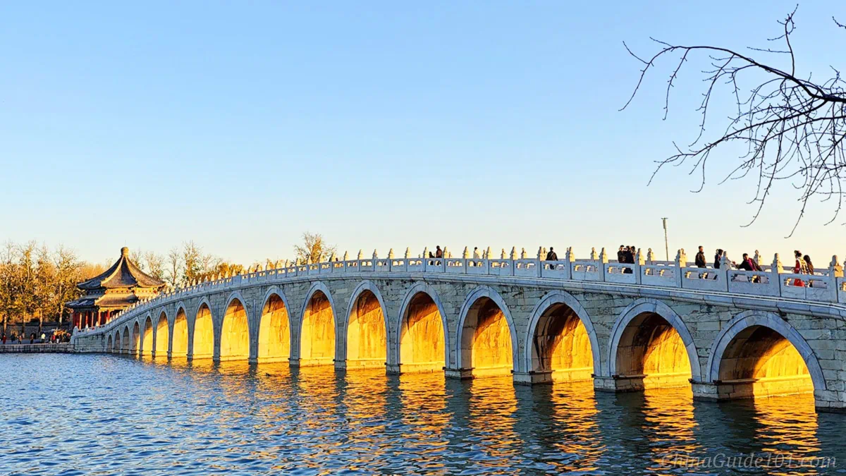 Low winter sunlight illuminating all 17 arches of the stone Seventeen-Arch Bridge, creating a golden glow underneath the arches reflected on Kunming Lake at the Summer Palace.