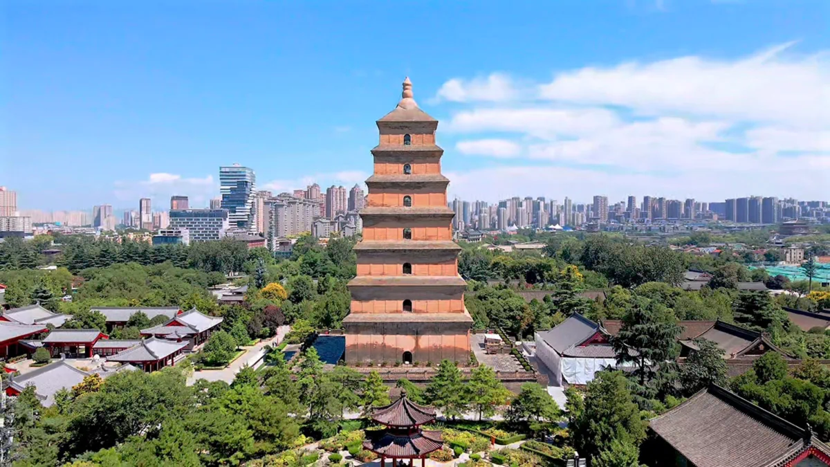 An aerial view of the ancient, multi-tiered Giant Wild Goose Pagoda in Xi'an, China, standing prominently against a clear blue sky. Traditional temple buildings with tiled roofs surround its base, amidst lush green trees. In the distance, a modern cityscape with high-rise buildings contrasts sharply with the ancient structure.