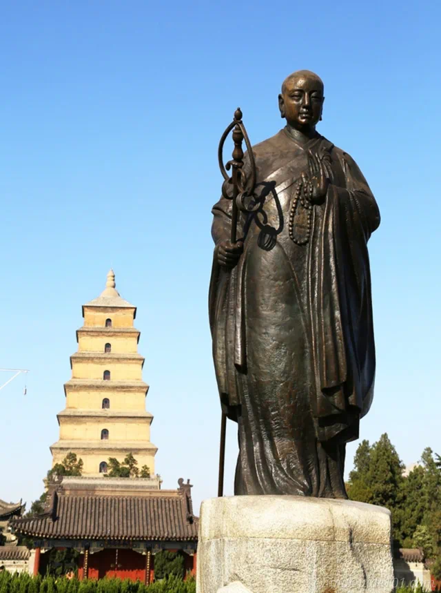 A tall bronze statue of the Tang Dynasty monk Xuanzang holding a staff and prayer beads, standing in the foreground with the historical Giant Wild Goose Pagoda and temple gate behind him under a clear blue sky.