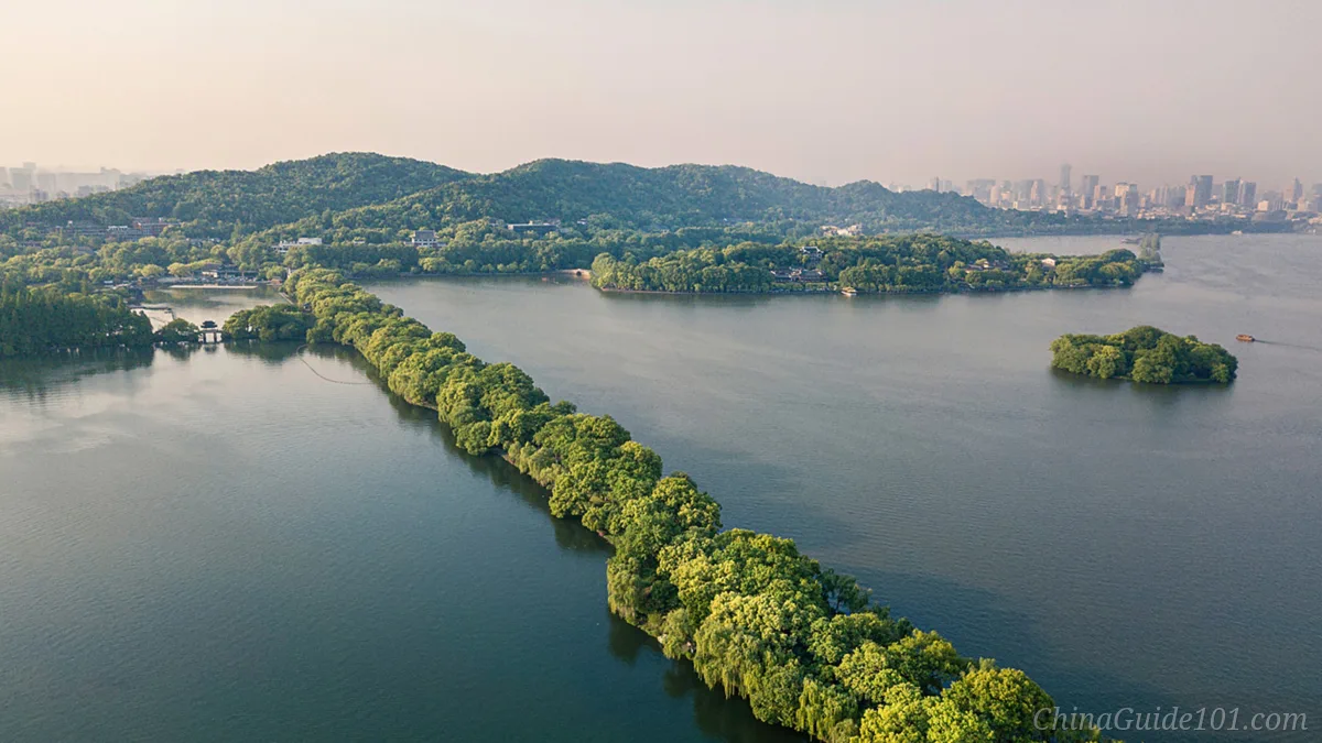 Aerial view of Su Causeway stretching across West Lake in Hangzhou, lined with lush green trees and surrounded by calm waters and mountains.