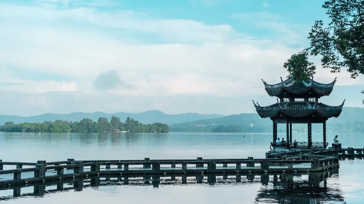Traditional Chinese pavilion on West Lake in Hangzhou with distant mountains and calm water.