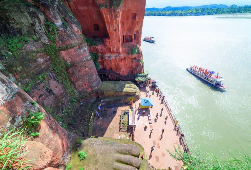 A high-angle view looking down at the giant feet of the Leshan Giant Buddha, the stone viewing platform below filled with tourists, and a cruise boat passing by on the river.