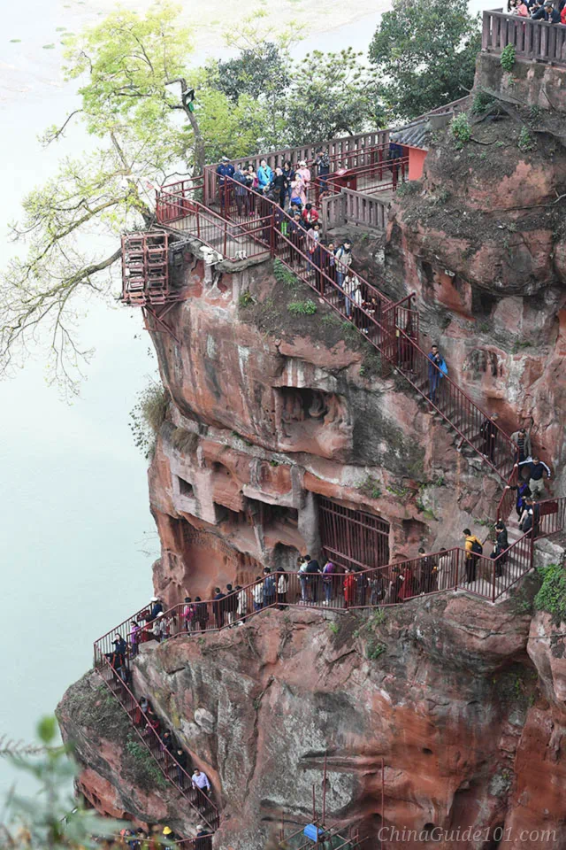 Visitors walking down a steep, narrow staircase path carved into the red cliff face beside the Leshan Giant Buddha in Sichuan, China.